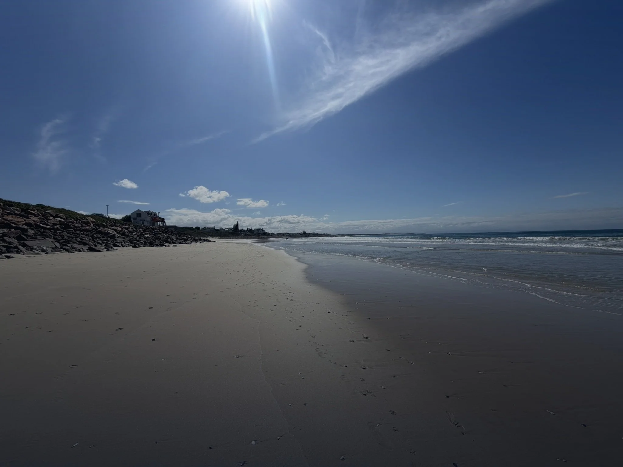 A sandy beach with footprints, rocks along the shore, a house with a deck and a windmill in the background, waves gently hitting the shore, and a clear sky with some clouds and the sun.