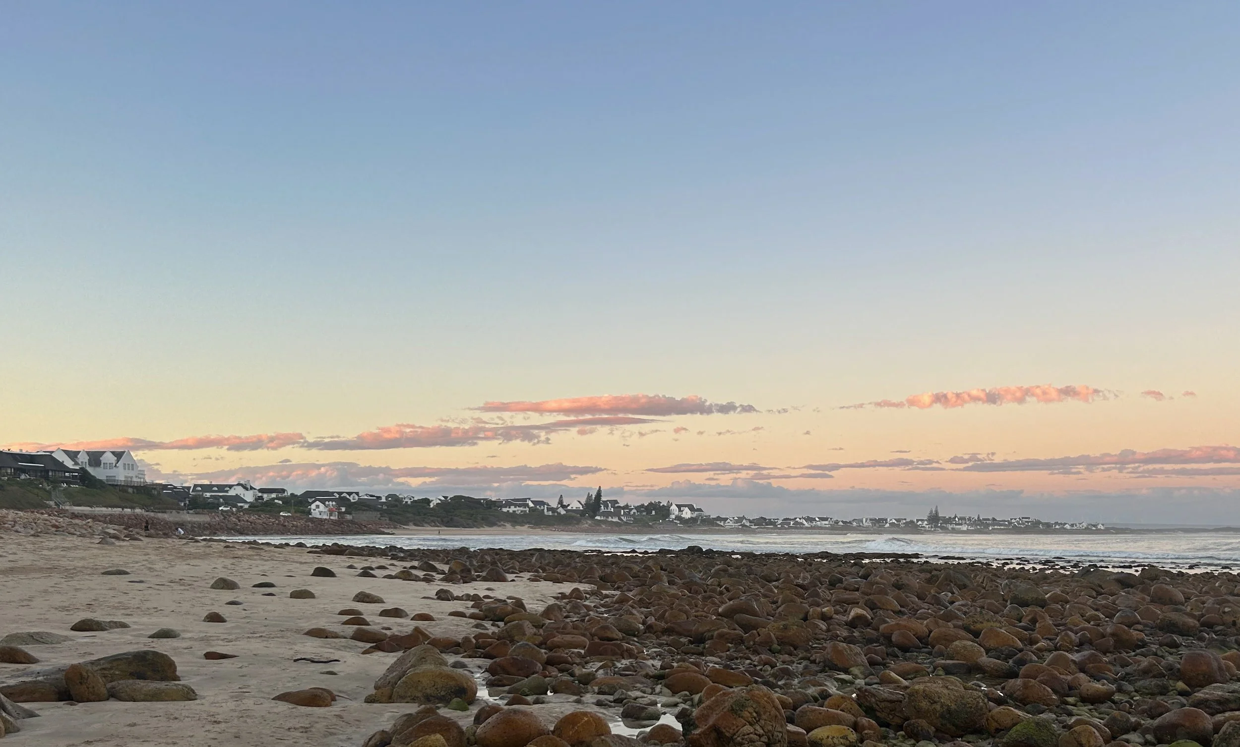 Scenic view of a beach with rocks, shoreline, and houses on a hill under a sky at sunset or sunrise.