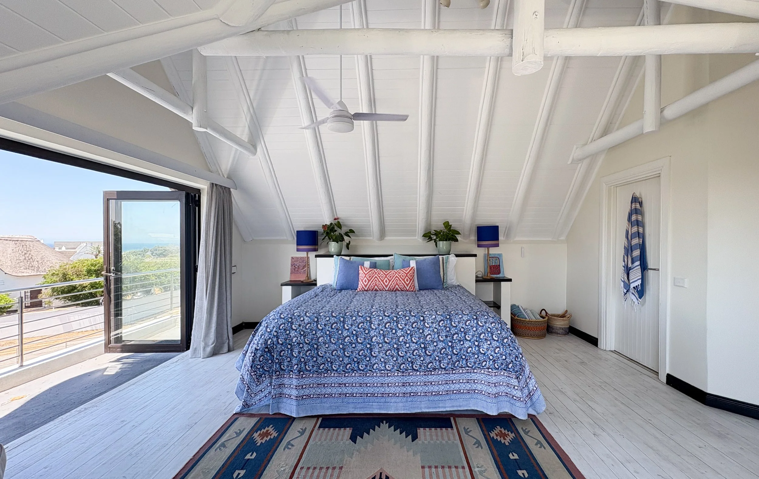 A bedroom with white wooden ceiling beams and a ceiling fan. There is a large window and a glass door that opens to a balcony. The bed has a blue patterned bedspread and various pillows. Two matching bedside tables with blue lamps and potted plants are on each side of the bed. There are woven baskets and a striped towel hanging on the door.