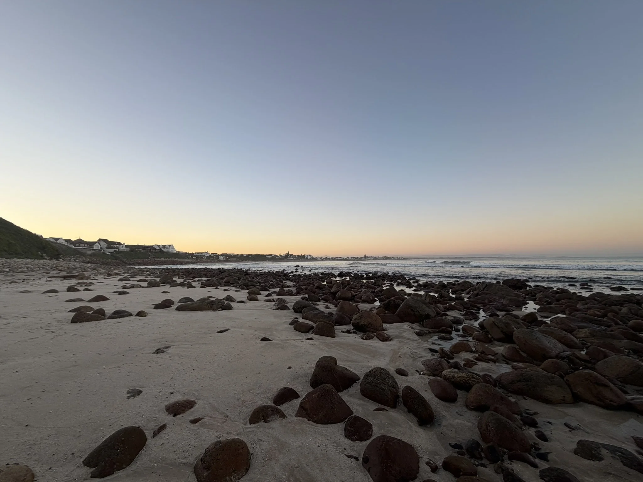 A beach at sunset with scattered rocks on the sandy shore, distant houses on a grassy hill, and calm ocean waves reaching the shore.