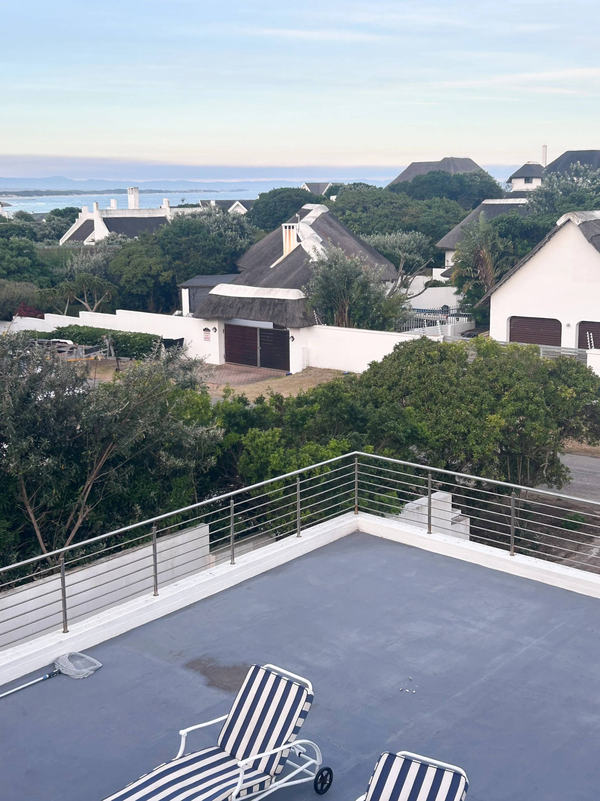 A rooftop patio with two striped lounge chairs, a small net, and a view of houses with thatched roofs, green trees, a fence, and the ocean in the background.