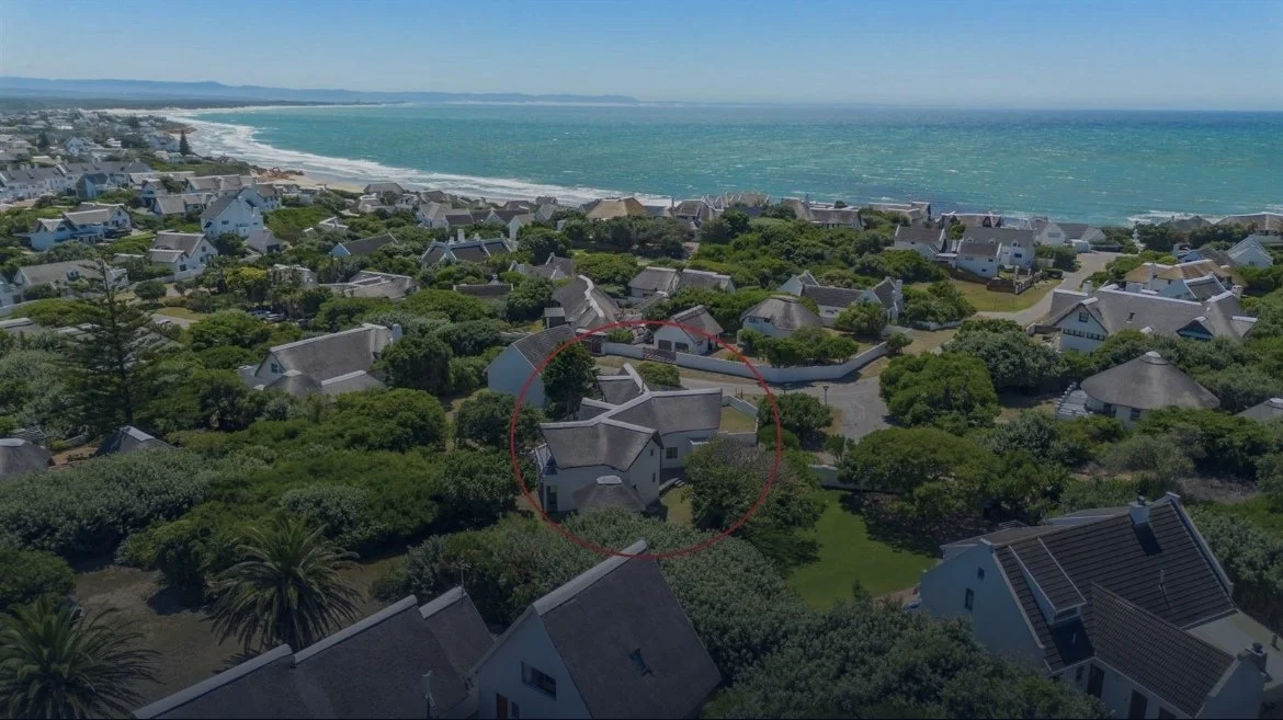 Aerial view of a coastal residential neighborhood with houses surrounded by greenery, ocean in the background, and a red circle highlighting a specific house.