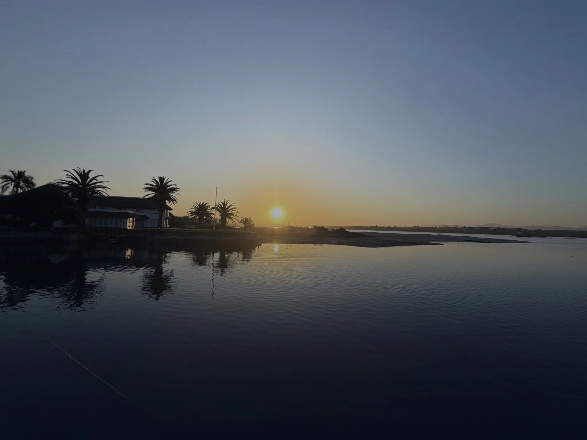 Sunset over a body of water with palm trees and a building in the foreground.