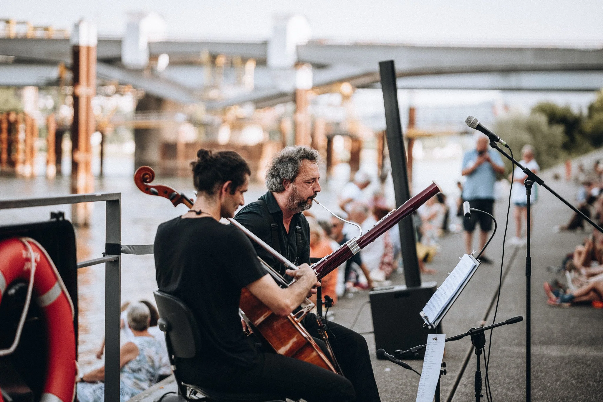 Zwei Musiker spielen auf einer Bühne im Freien, der eine mit einem Cello und der andere mit einer Querflöte, während im Hintergrund ein Publikum und eine Brücke zu sehen sind. Marvin Lahrs Musikevent Fotograf