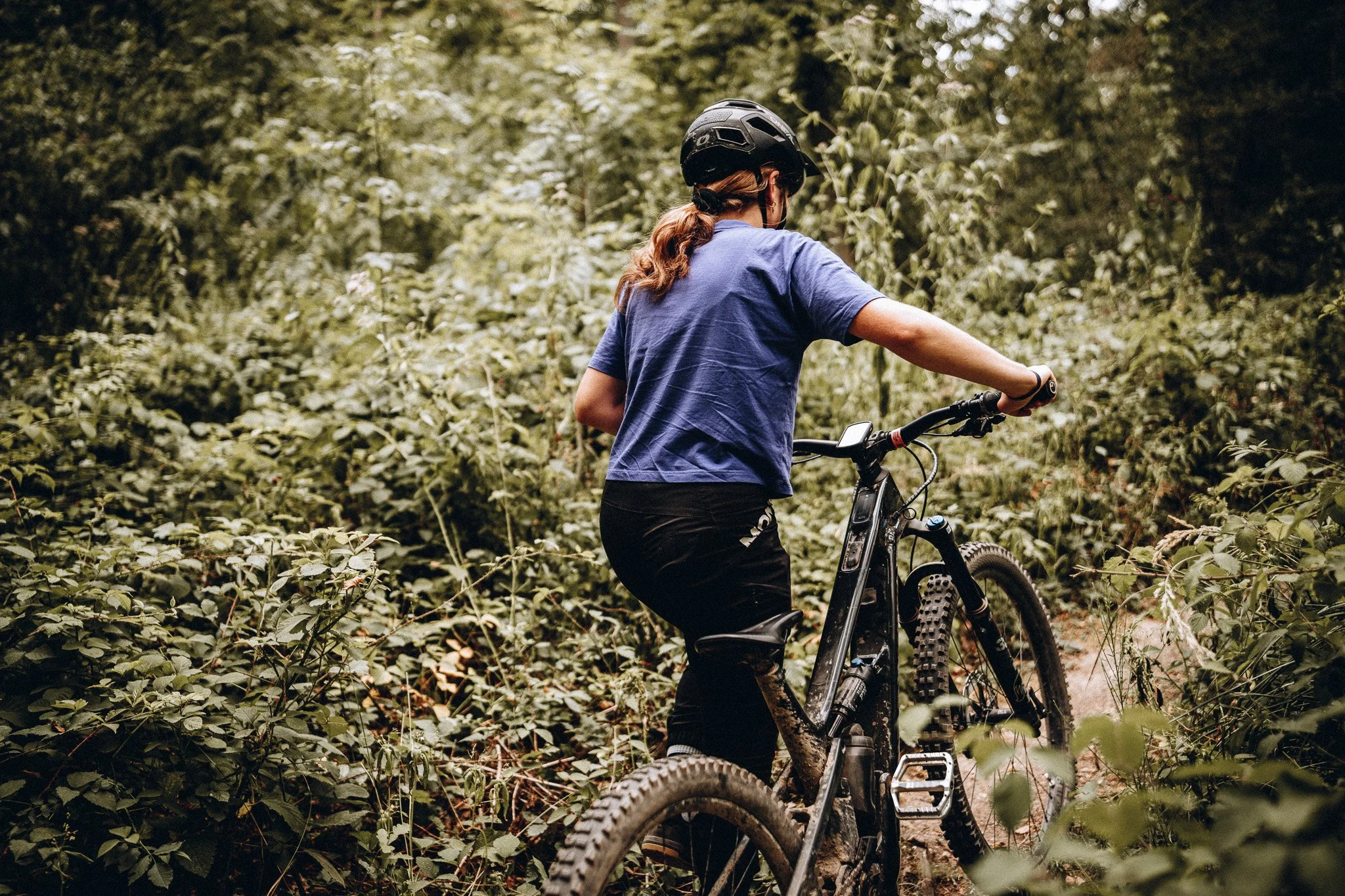 Frau mit Fahrrad in einem dichten grünen Wald, trägt Helm und blickt nach rechts. Marvin Lahrs Outdoor Mountainbike Fotograf aus Koblenz