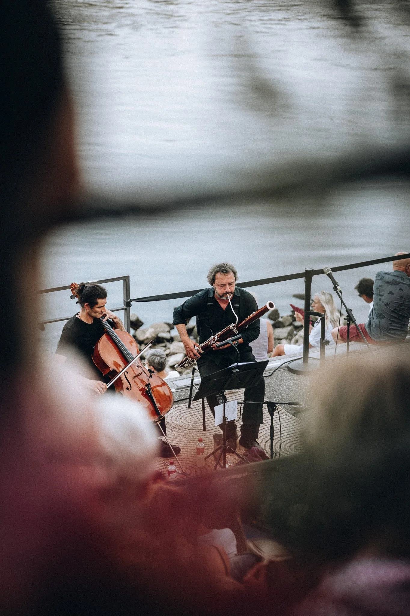 Musiker spielen an einem Ufer, im Vordergrund ein Cellist und ein Bassist, im Hintergrund Menschen, die Musik genießen, an einem See mit Steinen am Ufer.Marvin Lahrs Musikevent Fotograf Koblenz