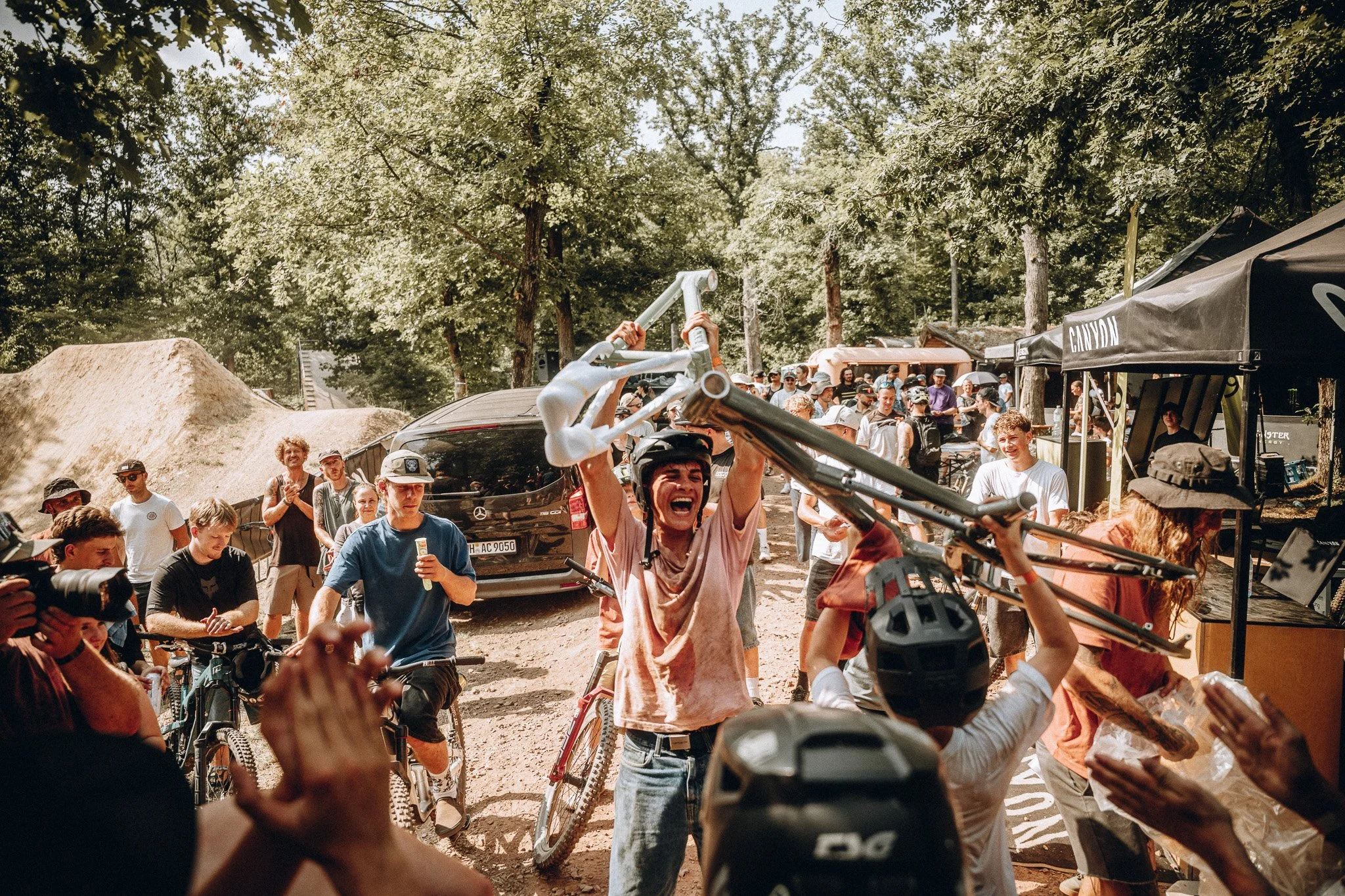 Menschen feiern bei einem Event in einem Wald, einige tragen Mountainbikes, im Hintergrund sind Bäume und einige Zelte. Marvin Lahrs Event Sport Fotograf