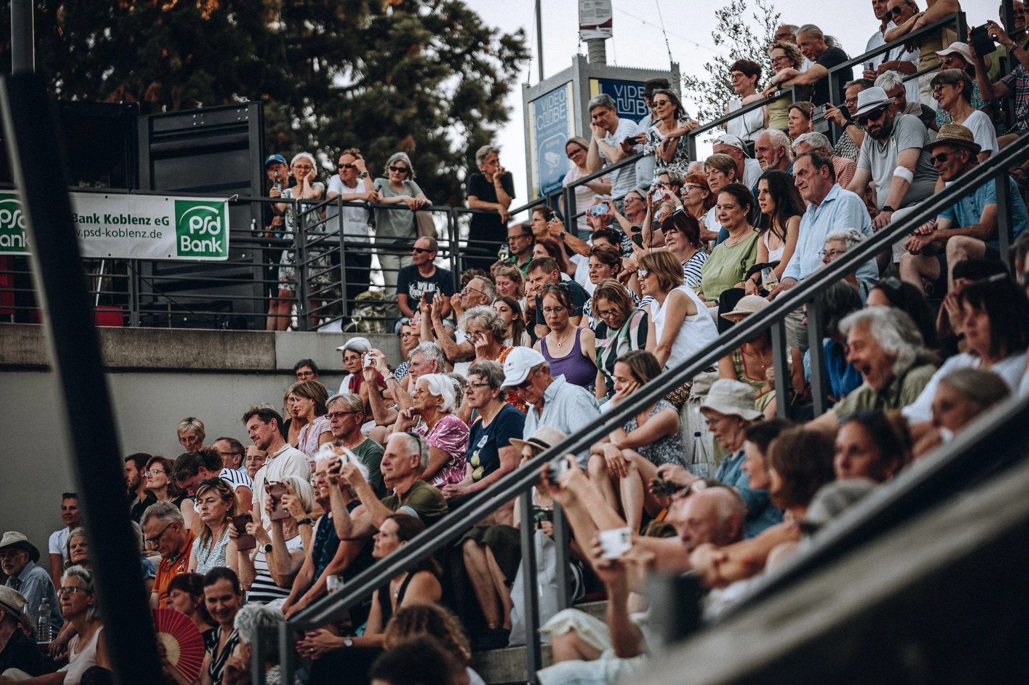 Menschen sitzen auf Tribünen bei einem Open-Air-Konzert oder Event im Freien, einige nehmen Fotos auf, während andere die Veranstaltung genießen. Marvin Lahrs Event Sport Fotograf Koblenz