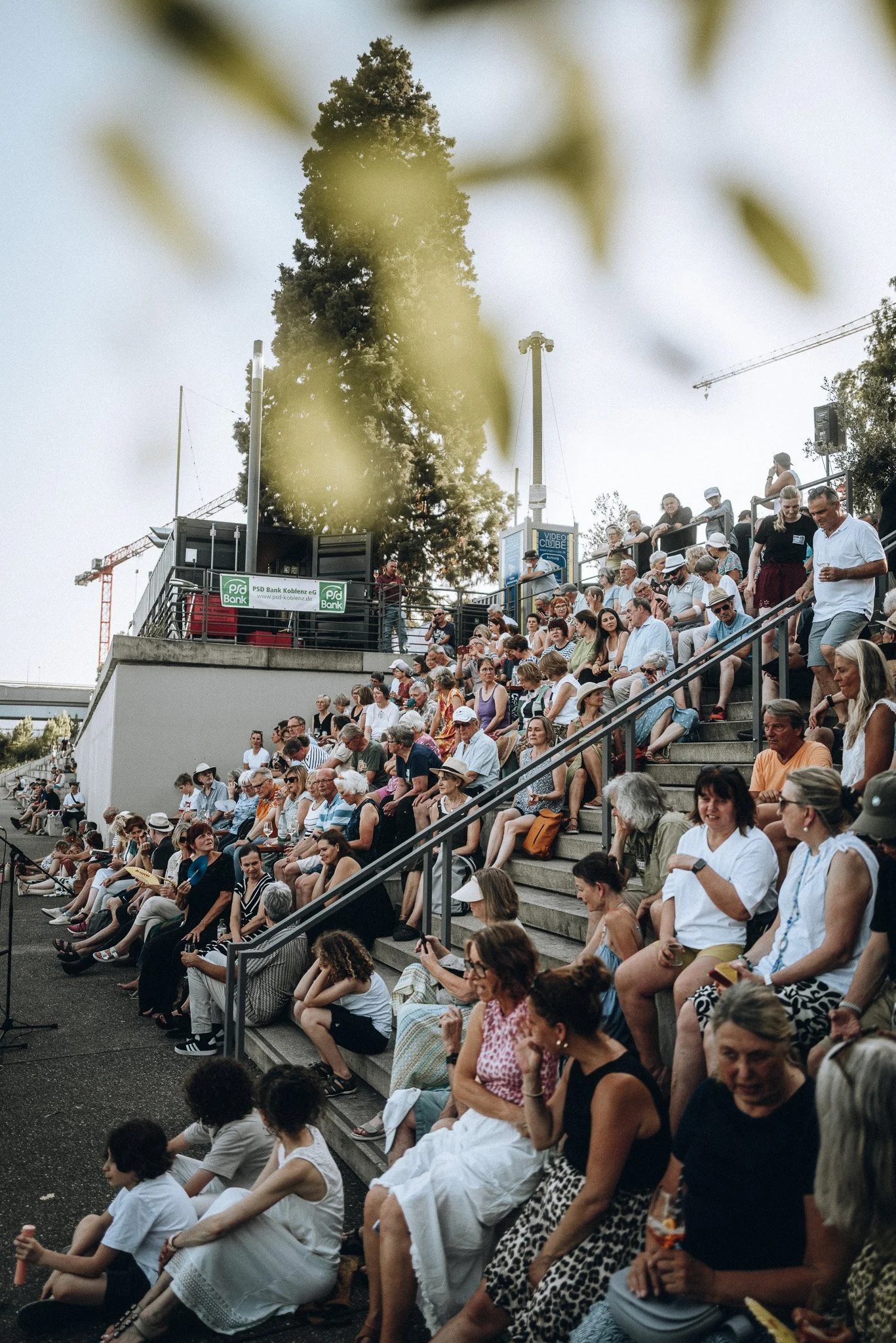 Menschen sitzen auf einer Freilufttribüne, einige trinken Getränke, andere schauen auf ein Ereignis. Im Hintergrund sind Bäume und Baugerüste sichtbar.Marvin Lahrs Musikevent Fotograf Koblenz