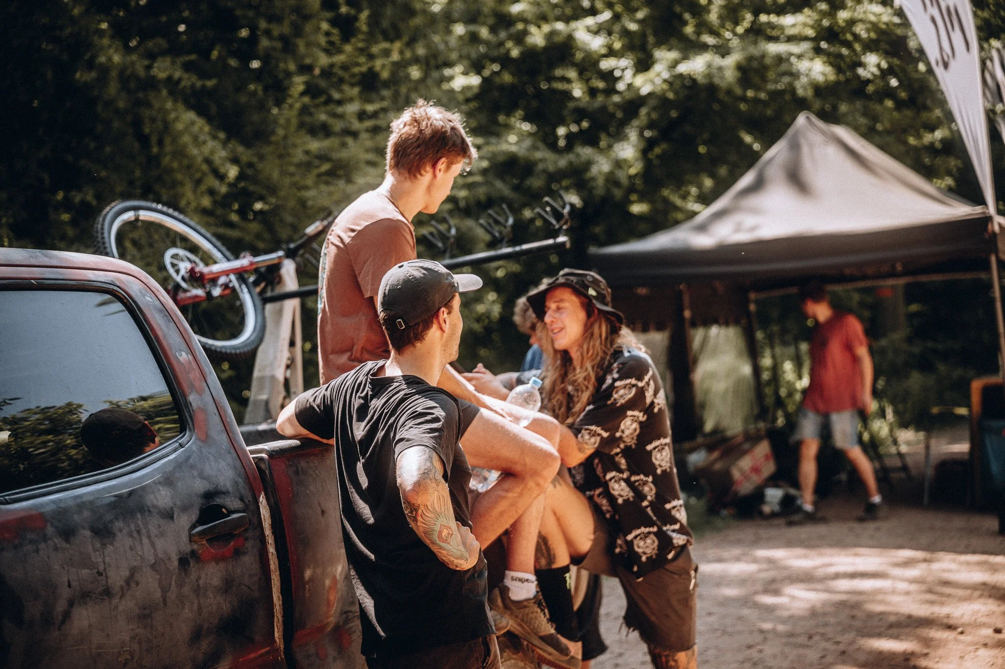 Gruppe von jungen Menschen unterhalten sich in einem Park. Ein Mann sitzt auf der Ladefläche eines Autos, während eine Frau und ein Mann stehen und sprechen. Im Hintergrund gibt es ein Zelt und Bäume.Marvin Lahrs Sport Fotograf