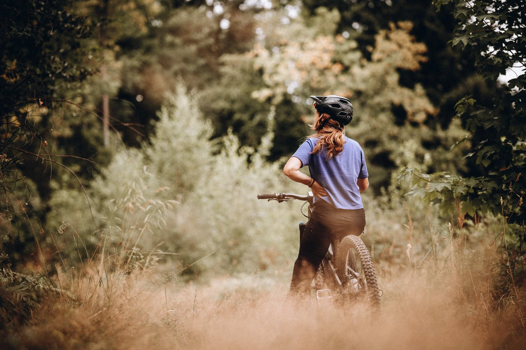 Eine Person mit einem Mountainbike steht in einem Wald und blickt in die Ferne. Die Person trägt einen schwarzen Helm und ein blaues T-Shirt und hat lange Haare. Marvin Lahrs Outdoor Fotograf aus Koblenz