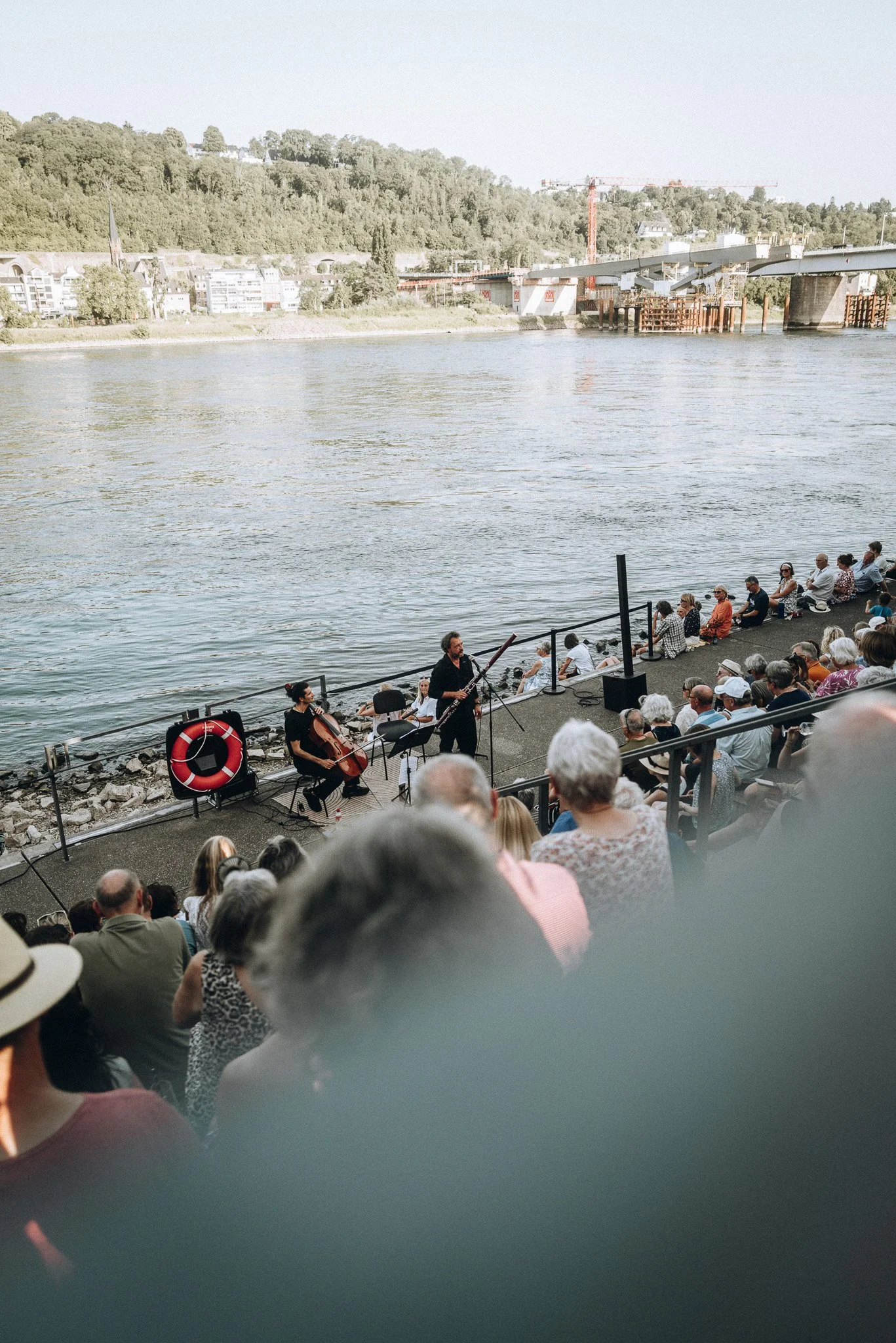 Menschen sitzen am Flussufer und hören einem Musiker zu, der mit einem Blasinstrument und einem Cellospiel auftritt. Im Hintergrund ist eine Brücke und eine Baustruktur sichtbar.Marvin Lahrs Musikevent Fotograf Koblenz