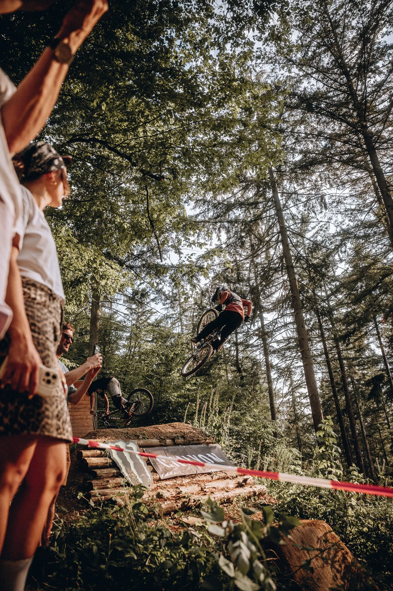 Mountainbiker springt in einem Waldparcours vor einer Zuschauermenge. Zuschauer fotografieren oder filmen, einige tragen Sonnenbrillen. Das Gelände ist aus Holzstämmen und Naturmaterialien gestaltet, in der Nähe liegt ein Banner mit einem Monster Ene