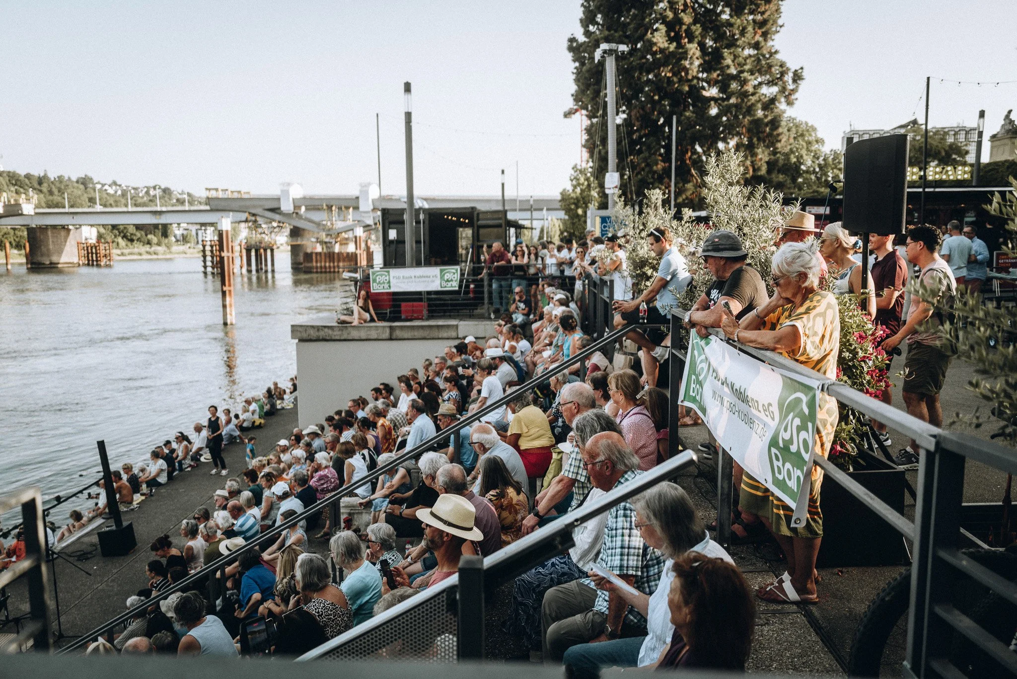 Menschen sitzen und stehen an einem Flussufer während eines Events, mit Bühnenaufbauten im Hintergrund und Bäumen entlang des Flusses. Marvin Lahrs Event Sport Fotograf Koblenz