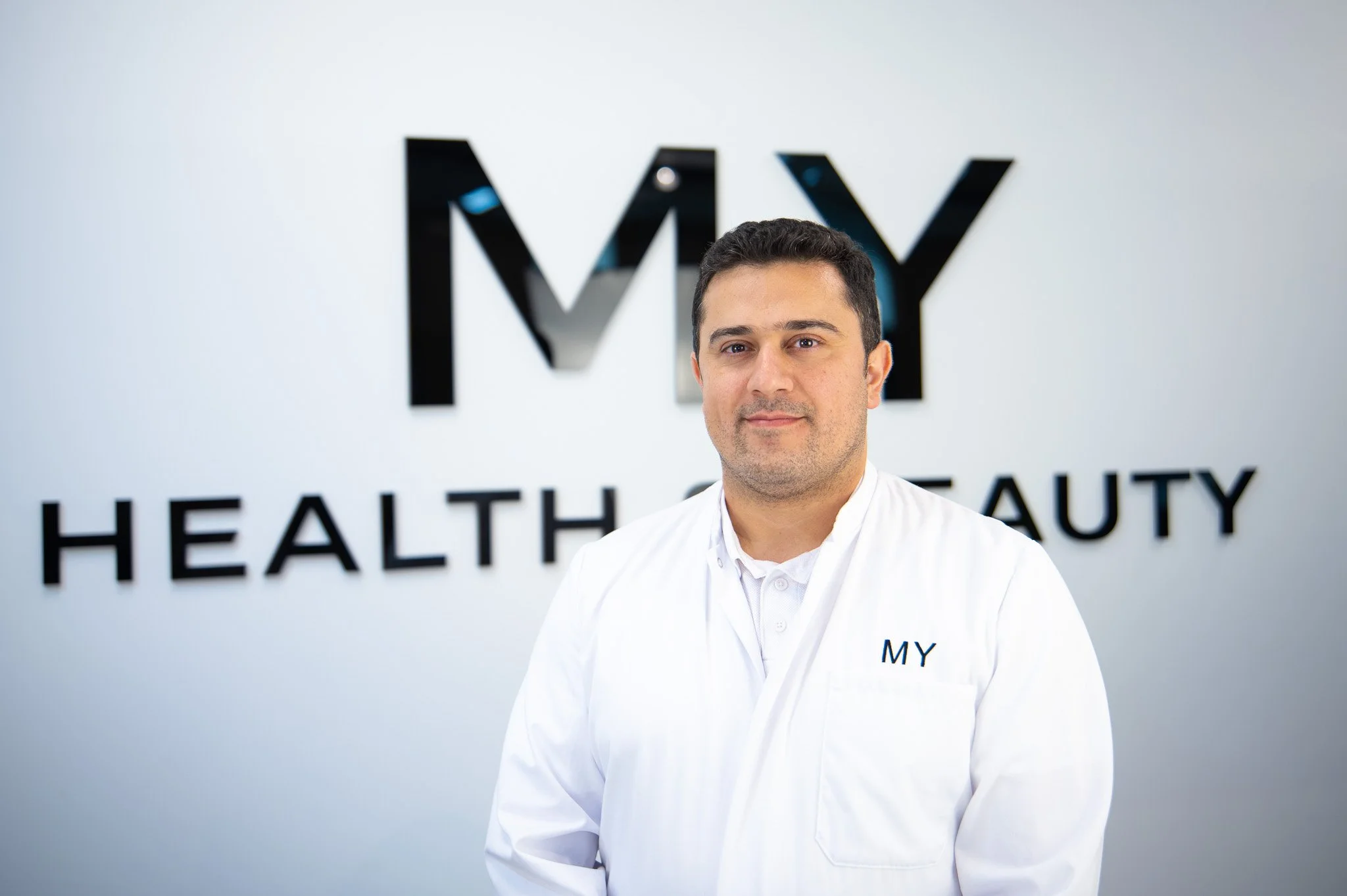 Ein Mann in einem weißen Arztkittel steht vor einer weißen Wand mit schwarzem Schriftzug, der das Wort 'MY' und einen Teil des Wortes 'HEALTH & BEAUTY' zeigt. Marvin Lahrs Businessfotograf aus Koblenz