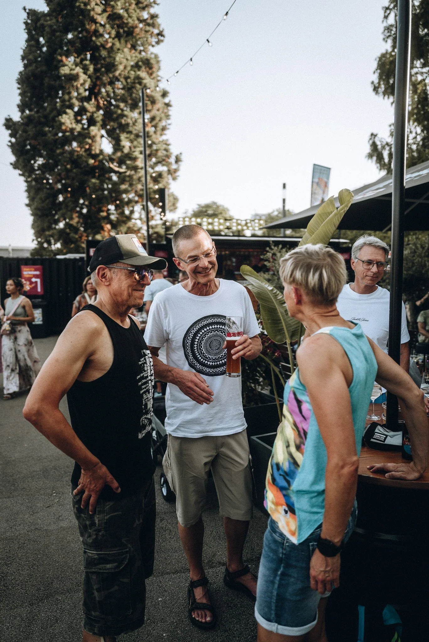 Eine Gruppe von vier Menschen, die sich im Freien an einem Abend unterhalten, während sie Getränke trinken. Es gibt Bäume im Hintergrund und einen Marktstand auf der rechten Seite.  Marvin Lahrs Event Sport Fotograf Koblenz