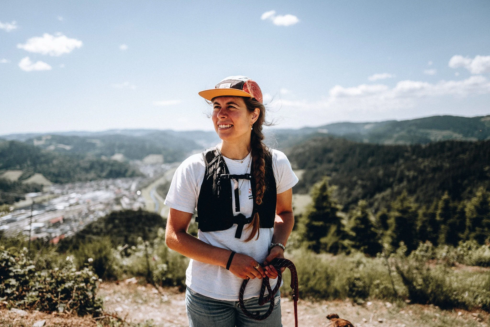 Junge Frau beim Wandern in den Bergen mit Kapuzenjacke und Rucksack, hält eine Leine, im Hintergrund flaches Tal und bewölkter Himmel. Zeigt Reise Fotografie