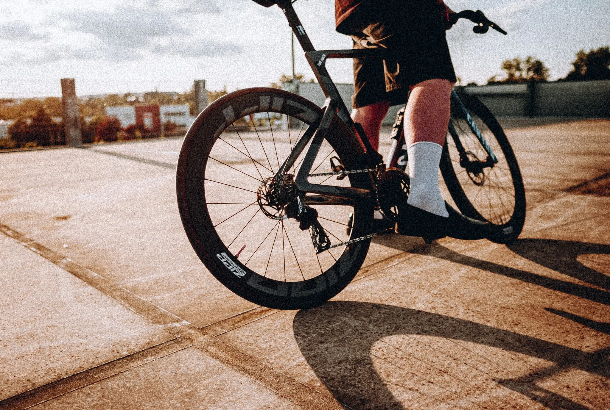 Eine Person auf einem schwarzen Fahrrad, trägt rote Shorts, schwarze Jacke und weiße Socken, auf einem offenen Platz bei Sonnenuntergang.