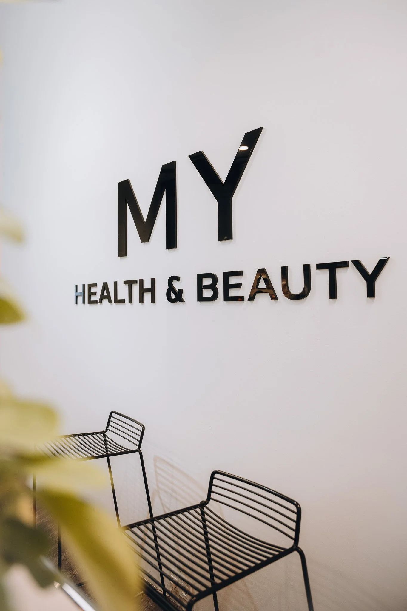 Schwarzweißes Regal in einem Raum, an der Wand hängt ein großes schwarzes Schriftzeichen "MY" und darunter steht in Großbuchstaben "HEALTH & BEAUTY". Im Vordergrund sind zwei schwarze DrahtStühle sichtbar, einer ist nur halb sichtbar. Marvin Lahrs 