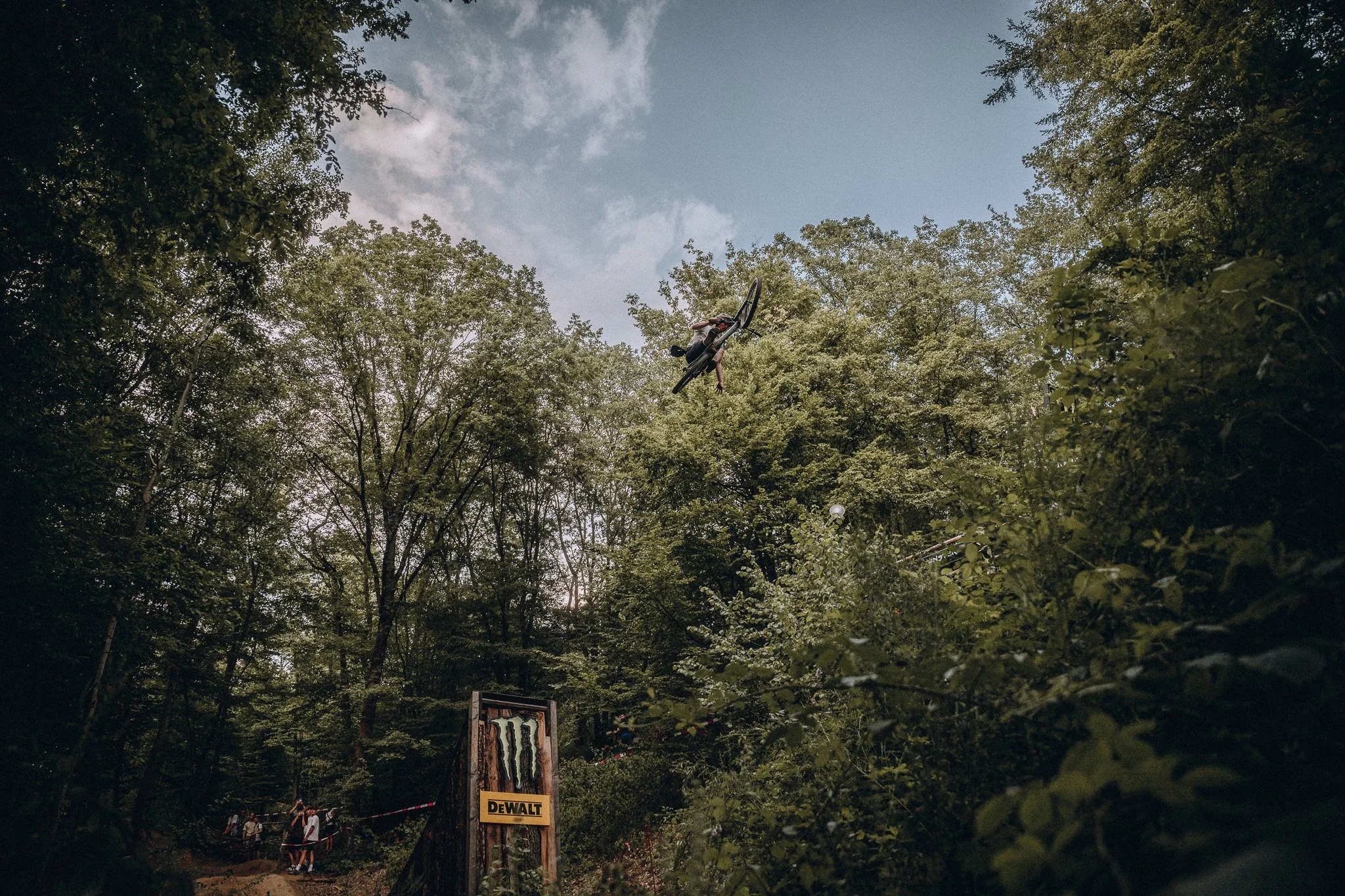 Ein Mountainbiker macht einen Sprung in einem Wald, der Himmel ist sichtbar durch die Baumkronen, im Vordergrund sind Besucher und Werbetafeln. Marvin Lahrs Sport Mountainbike Fotograf