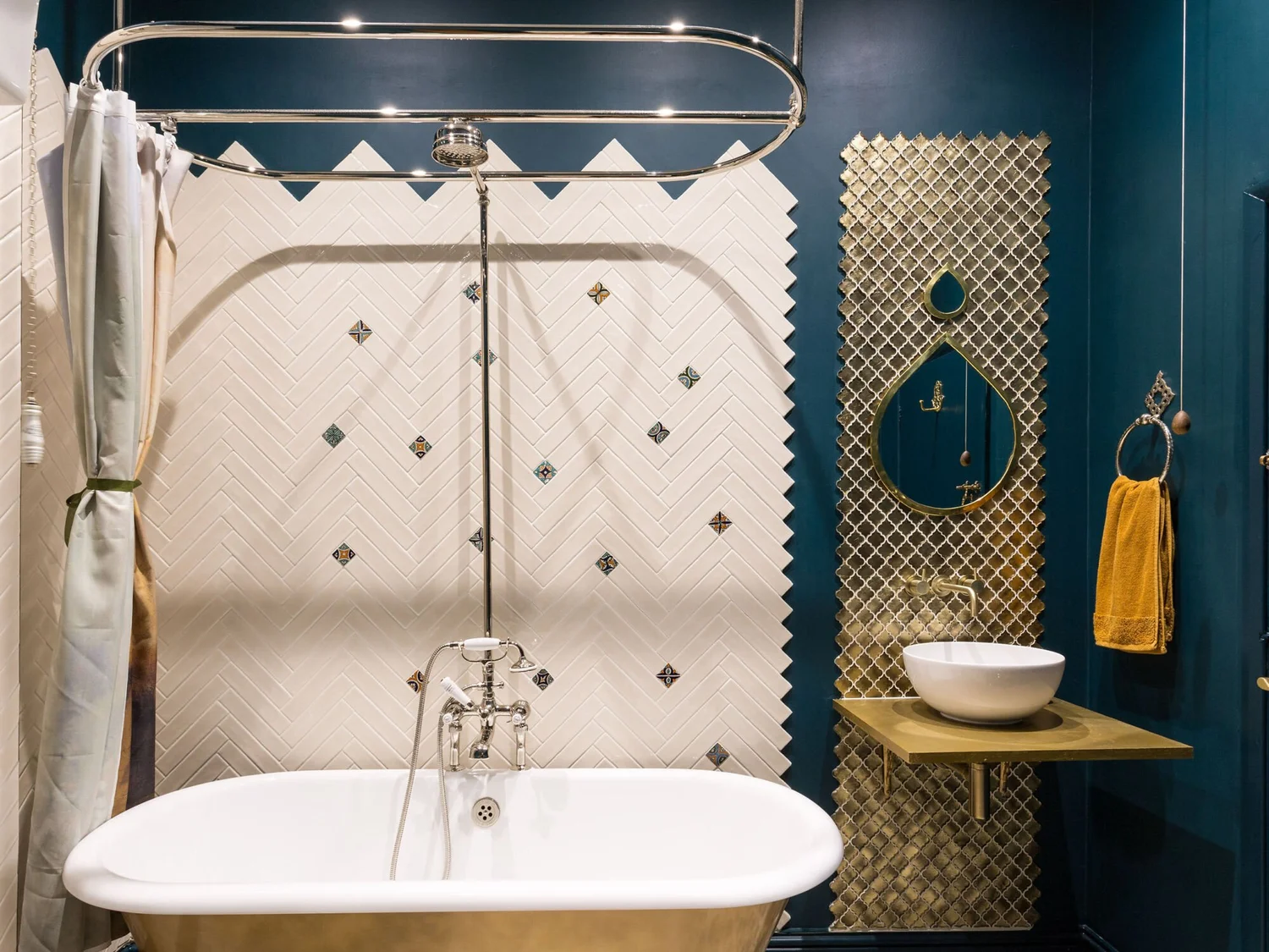 Bathroom with white herringbone shower tiles, dark teal walls, a freestanding bath and decorative gold tiles behind the basin.