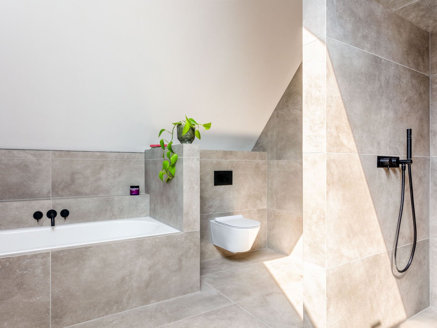 Modern bathroom with beige tiled walls, a white bathtub, a toilet, and a black showerhead. A green potted plant and a small purple container are on the ledge.