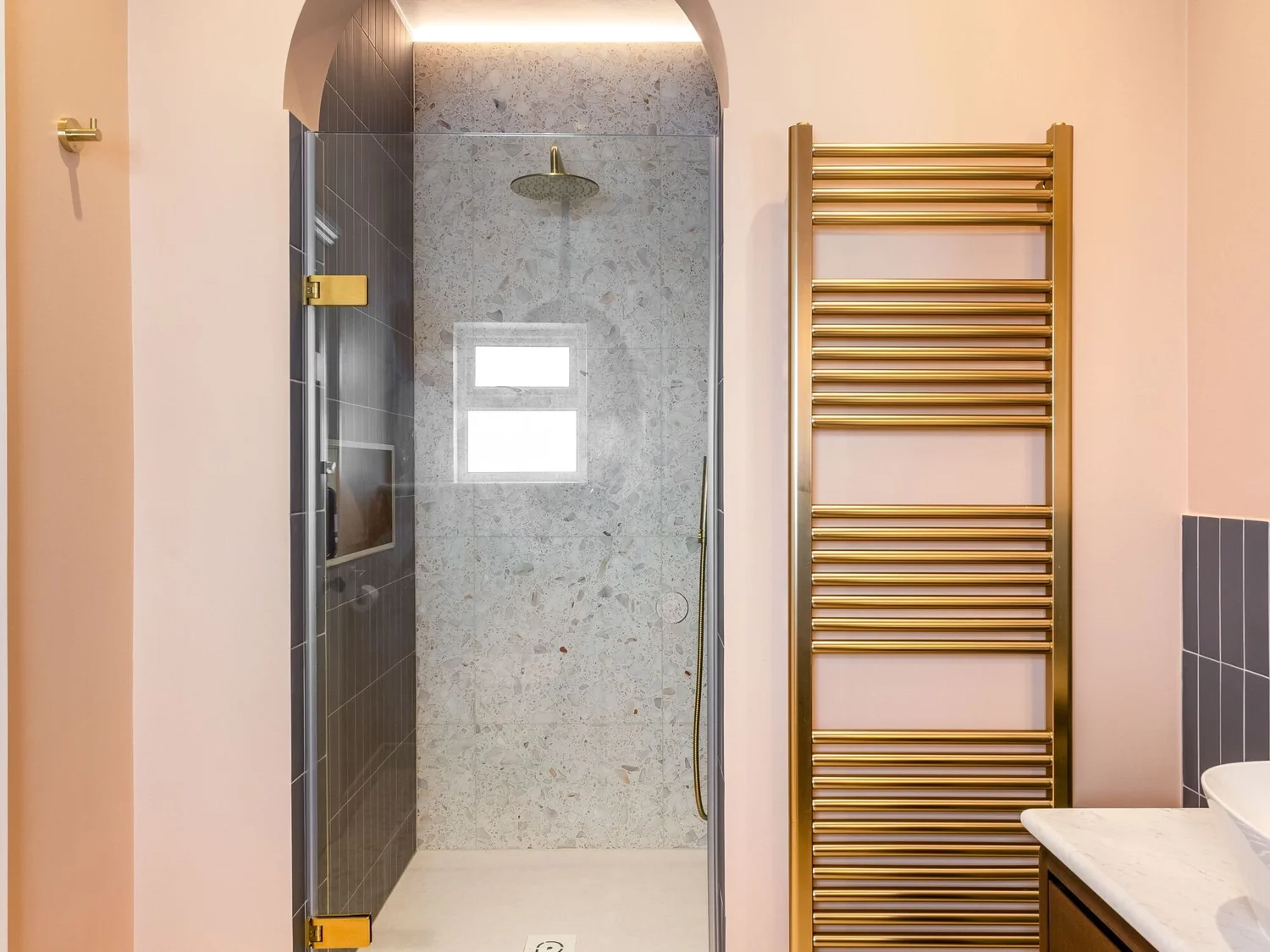 Walk-in shower with grey tiled walls, a brass rainfall shower, clear glass enclosure and gold heated towel rail beside a soft pink wall.
