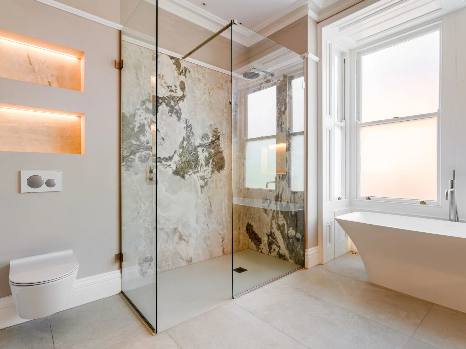 Bathroom with a glass shower enclosure, large marble-effect wall slabs, pale floor tiles and a freestanding bath.