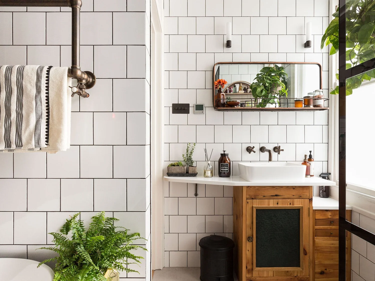 A modern bathroom with white subway tiles, a black metal pipe towel bar with a striped towel, a large mirror, a teak sink vanity, a white sink, a small potted plant, a black trash can, and a wall-mounted mirror with plants reflected in it.