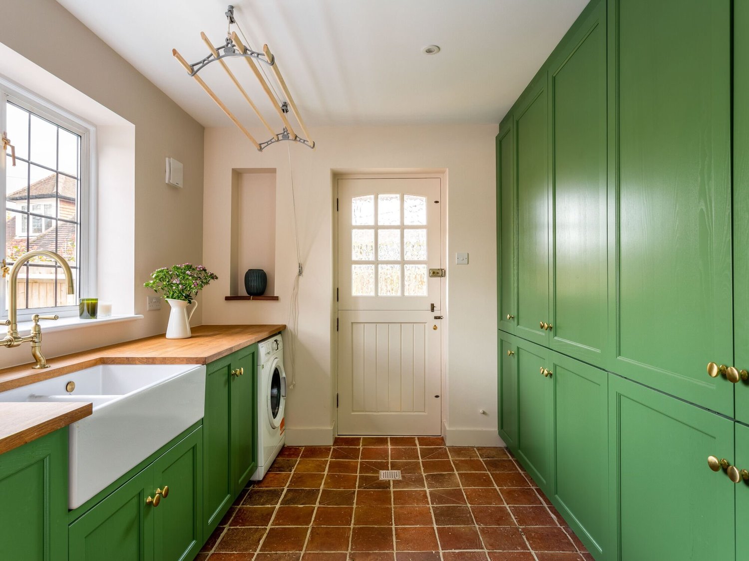 Utility room with green fitted cupboards, timber worktops, a white sink, washing machine and terracotta tiled floor.
