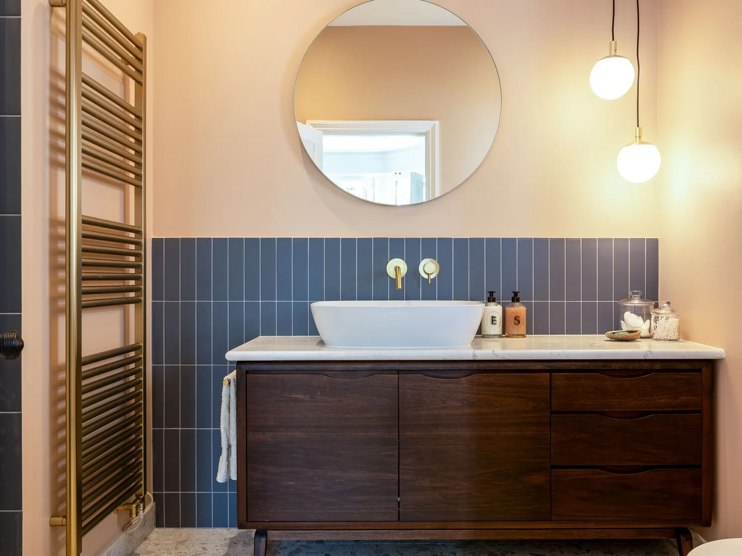 Bathroom vanity with a vessel sink, a round mirror, blue tiled backsplash, and two pendant lights. There is a wooden cabinet below the sink, toiletries on the counter, and a towel hanging on a brass towel bar. The wall is painted beige.