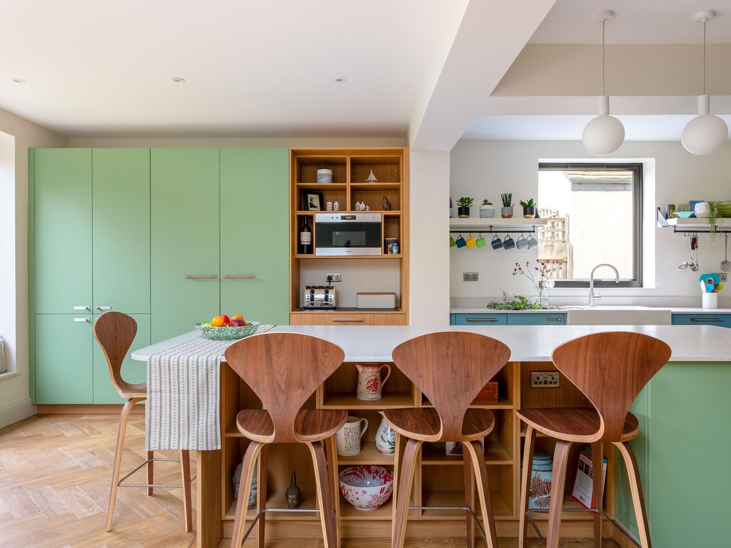 Green fitted kitchen with a white island worktop, open timber shelving and wooden bar stools.