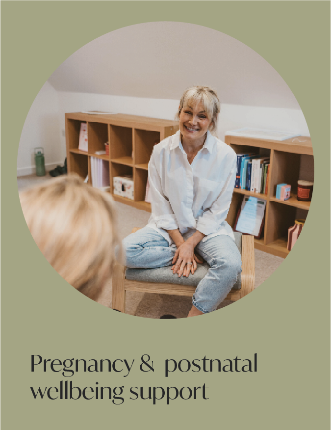 A smiling woman sitting on a chair in a room with bookshelves, talking to another person, promoting pregnancy and postnatal wellbeing support.