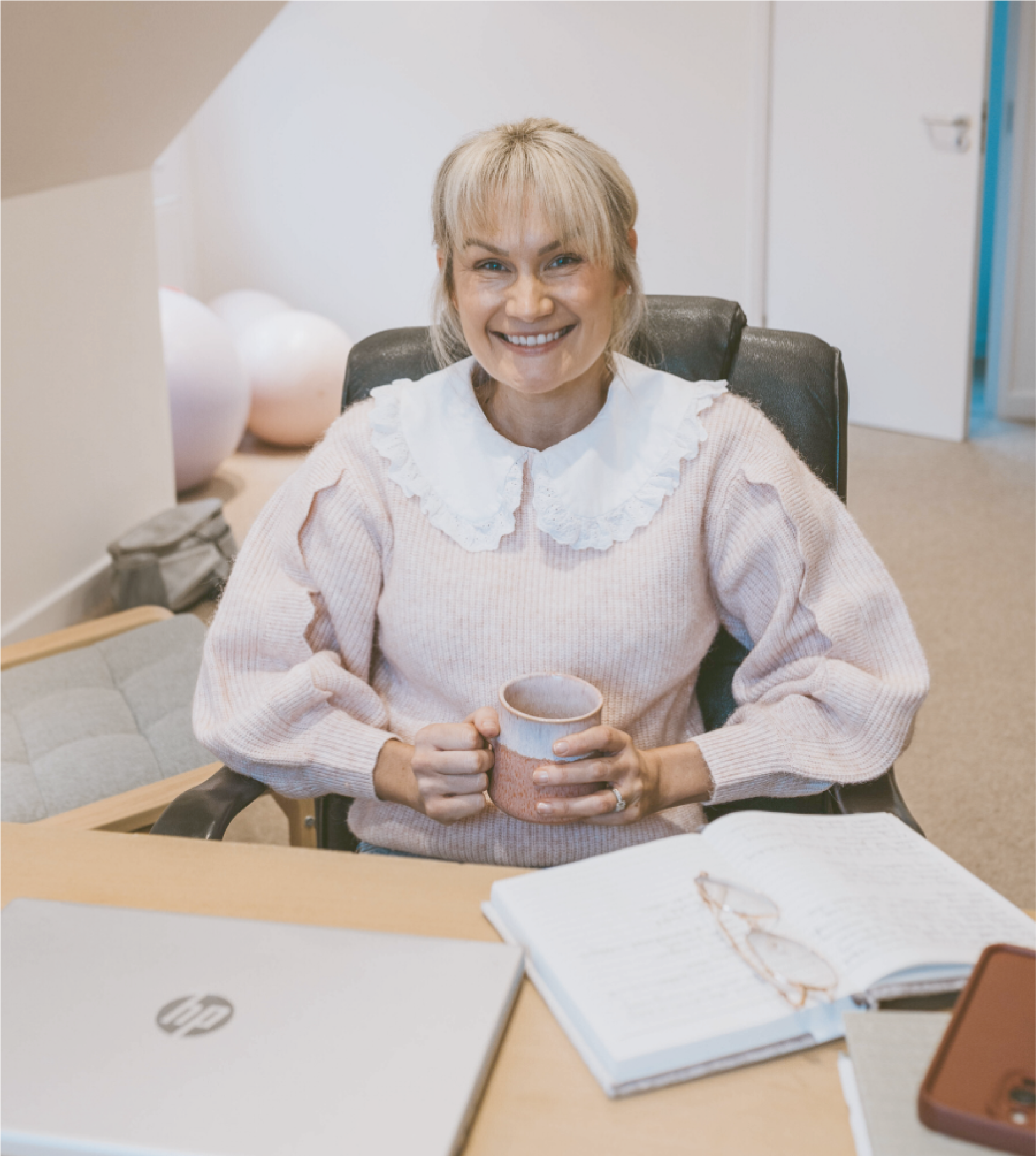 A woman smiling and sitting at a desk holding a coffee mug, with a cluttered desk and a notebook open in the foreground.