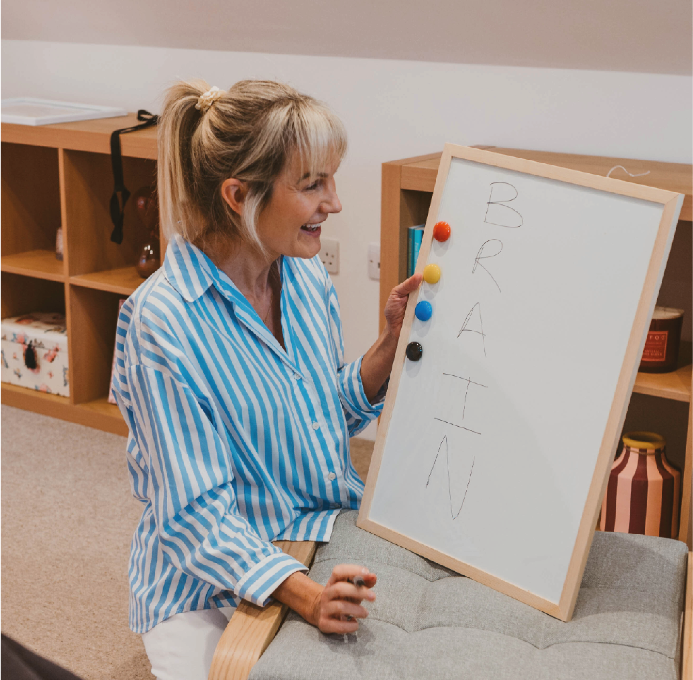 Woman with blonde hair in a ponytail wearing a blue and white striped shirt, smiling, holding a marker, standing next to a whiteboard with the alphabet written on it and colorful magnets, in a room with wooden shelves and a beige carpet.