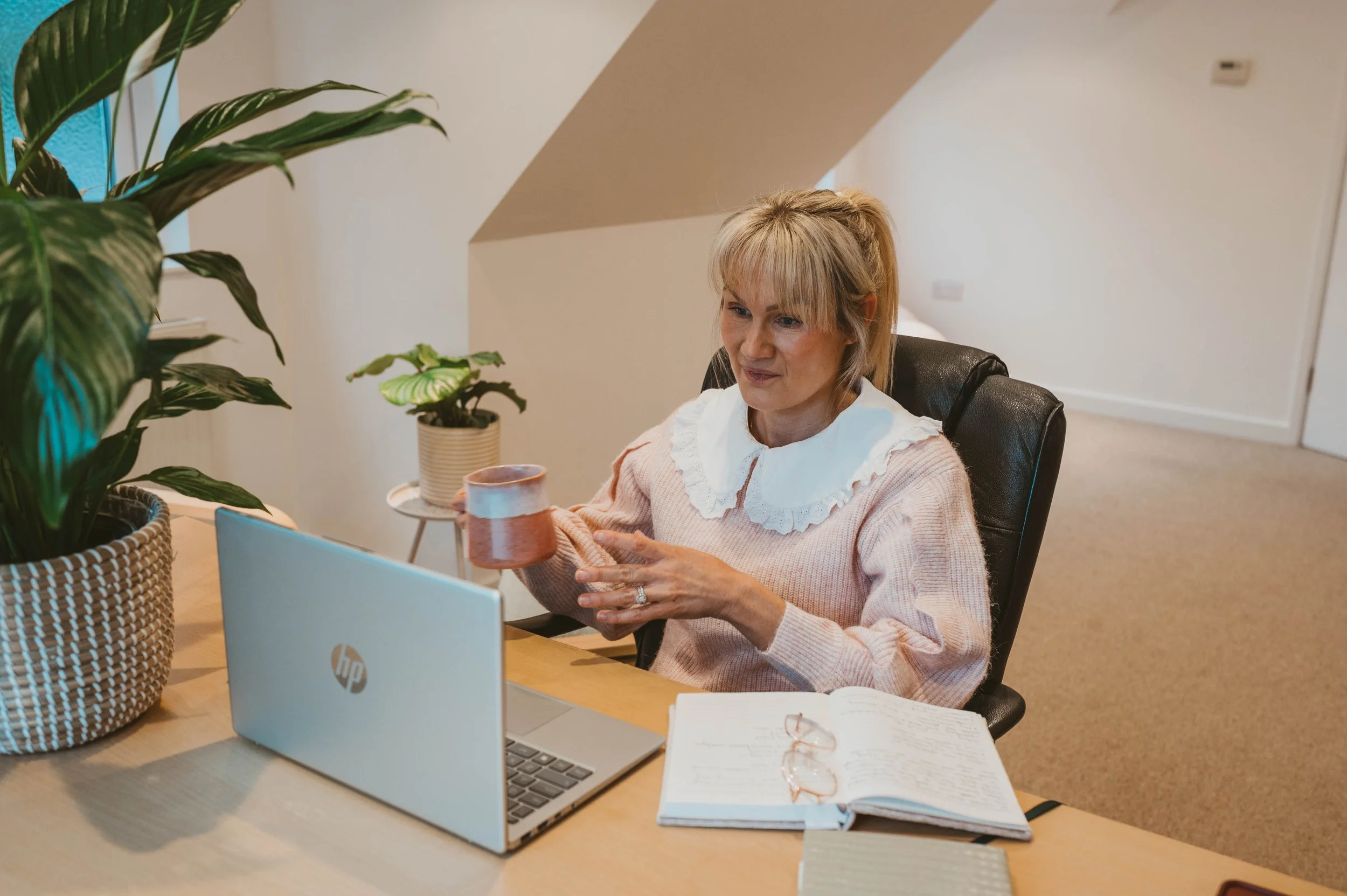A woman sitting at a desk in a home office, holding a coffee mug, with open notebook, glasses, laptop, and green potted plants.