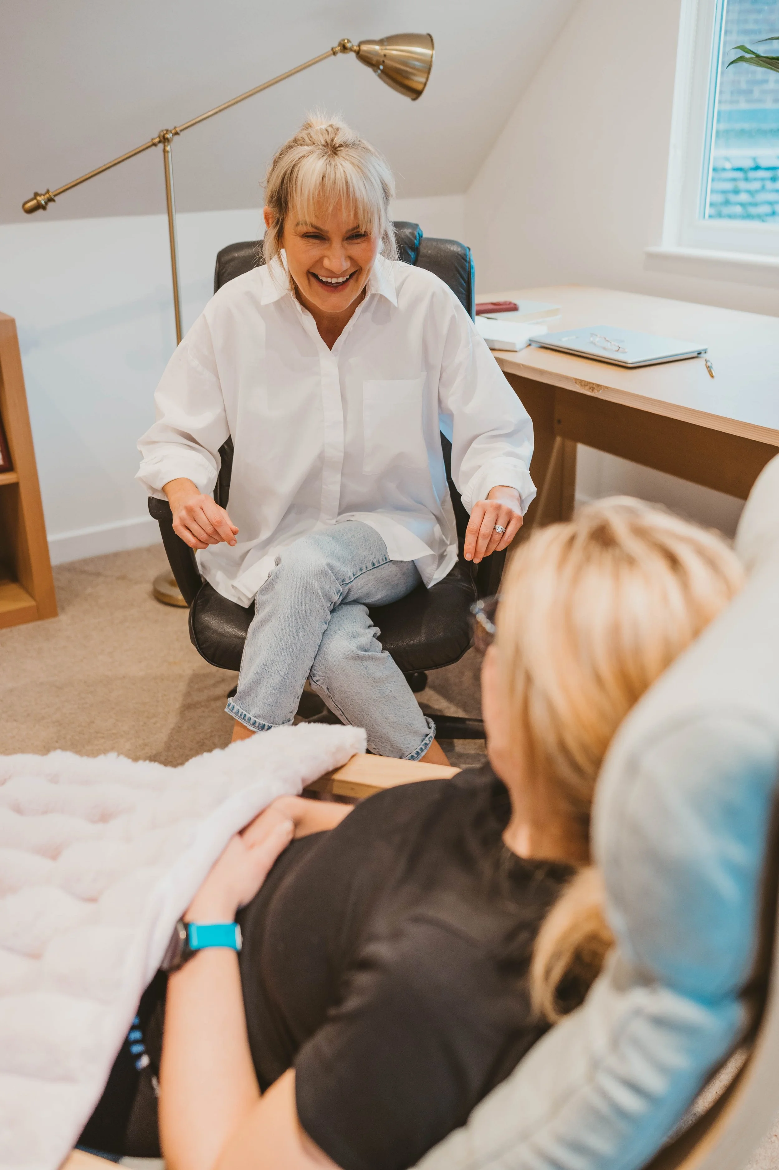 A woman in a white shirt sitting on a black office chair, laughing with a woman in black shirt and glasses, lying in bed with a blanket and wearing a blue wristband, in a cozy room with a desk, books, and a window.