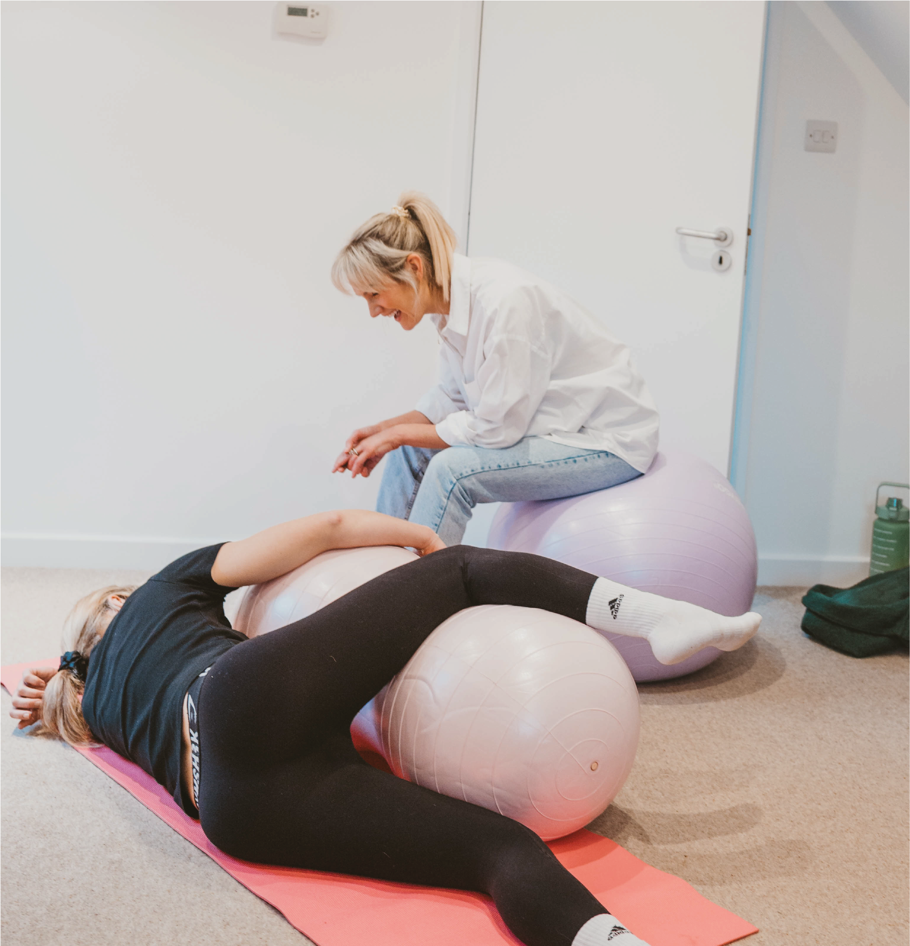 A woman lying on her side on a yoga mat with her head resting on her arm and her legs bent, stretching her back over a large pink exercise ball, while another woman sitting on the ball and leaning forward looks on and smiles.