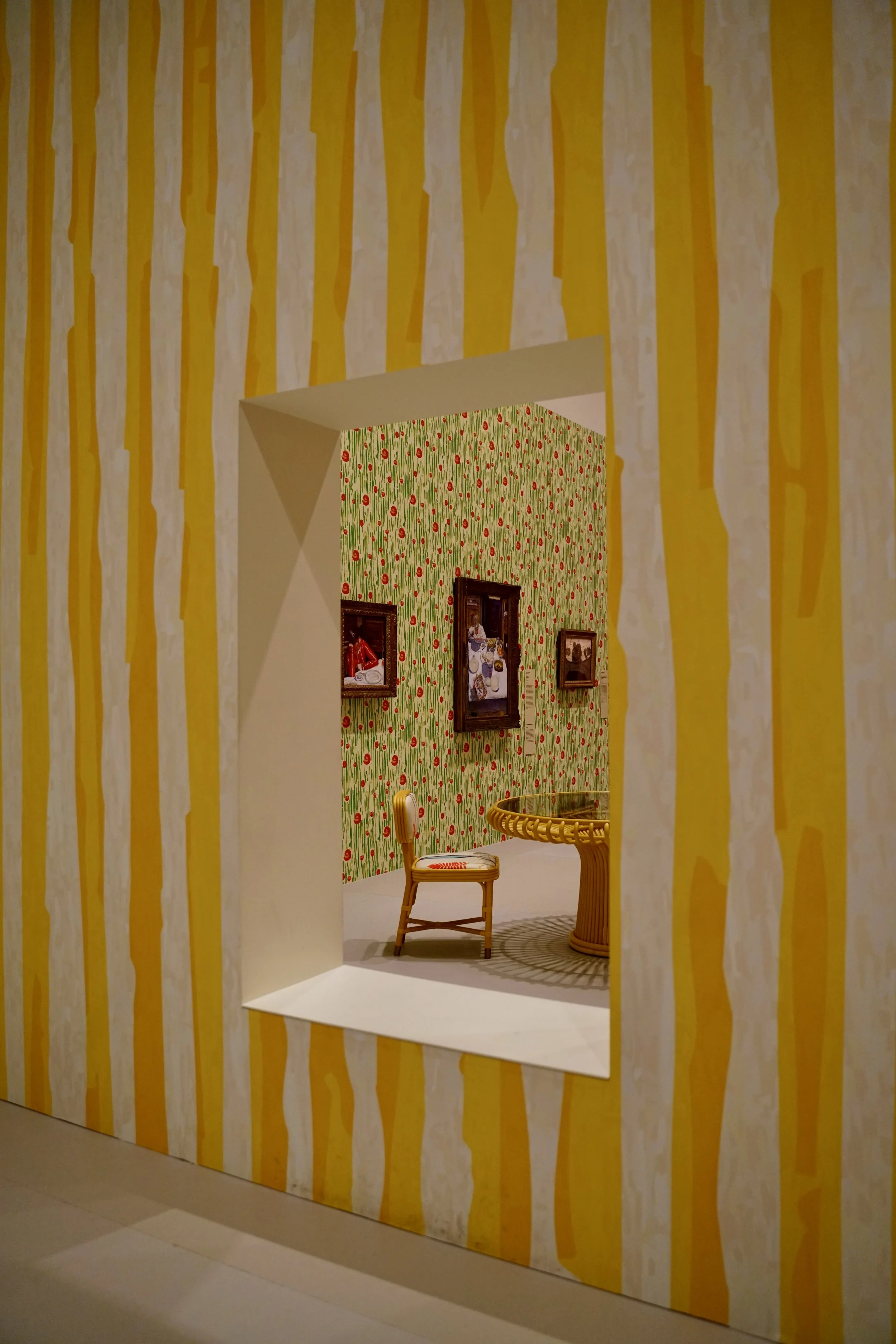 View through a window into a room with yellow and white striped wallpaper, a red and white patterned wall, framed pictures, a rattan table, and a chair. NGV photo: Brook Powell