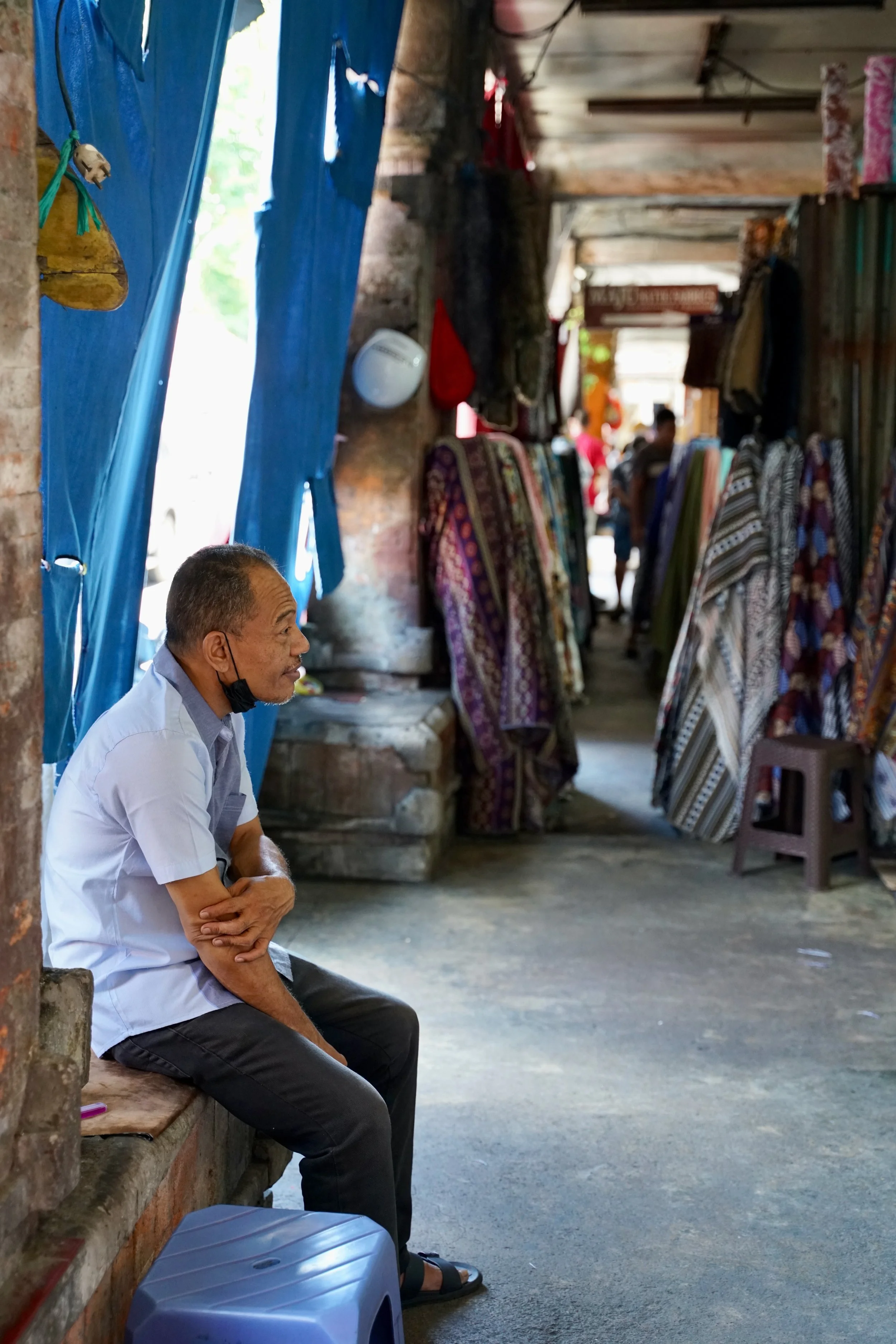 A man with short hair, wearing a white shirt and black pants, sitting on a brick bench inside a rustic shop. Cloth items are displayed on racks along the wall. The man has a black face mask pulled down below his chin, and a blue plastic container is on the floor near him. The shop has an open entrance with blue tarps hanging and natural light filtering in.