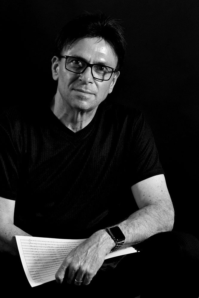 Black and white portrait of a man wearing glasses, a dark t-shirt, holding a sheet of music, and looking at the camera.