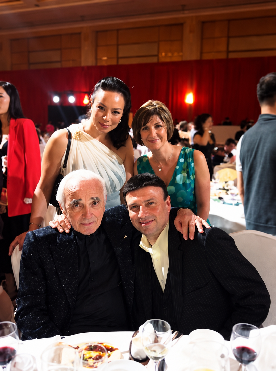 Group of five adults at a formal event, with two women standing behind two men seated at a banquet table, in a large hall with red curtains and dim lighting.