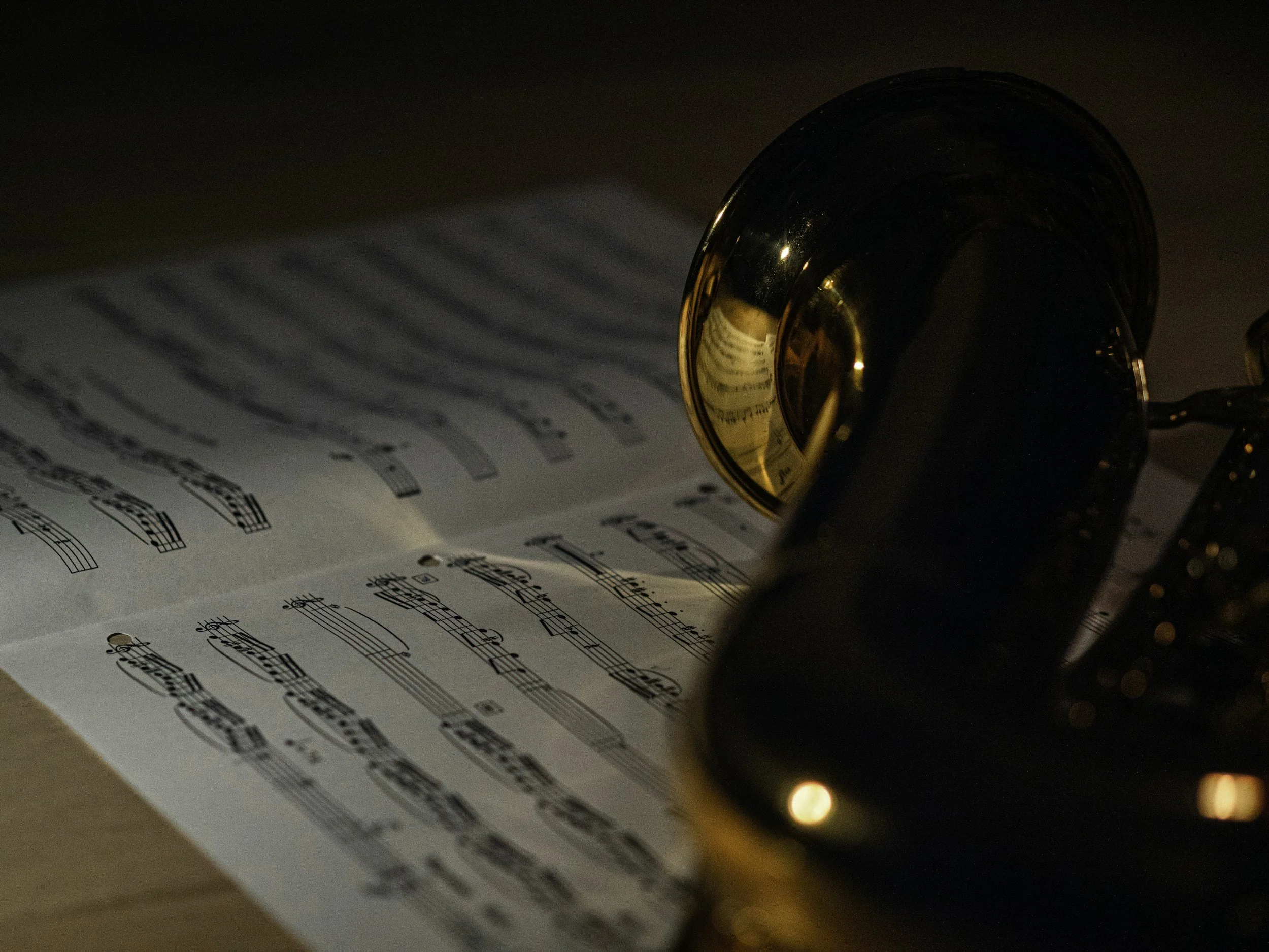 Sheet of printed sheet music with musical notes on white paper, placed under a shiny brass or gold-colored bell, possibly of a musical instrument or a decorative item, in a dimly lit setting with warm lighting reflections.