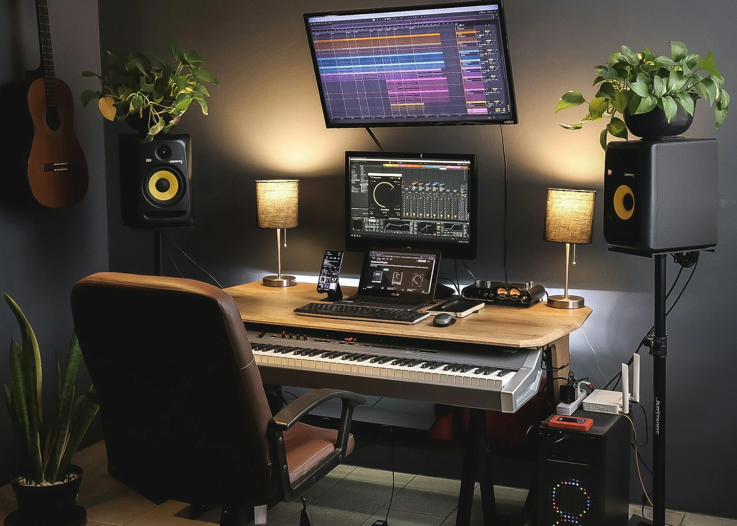 A music production workspace with a digital audio workstation on a large monitor, headphones, studio speakers, a MIDI keyboard, a laptop, a smartphone, two table lamps, two potted plants, a guitar on the wall, and various audio equipment in a dimly lit room.