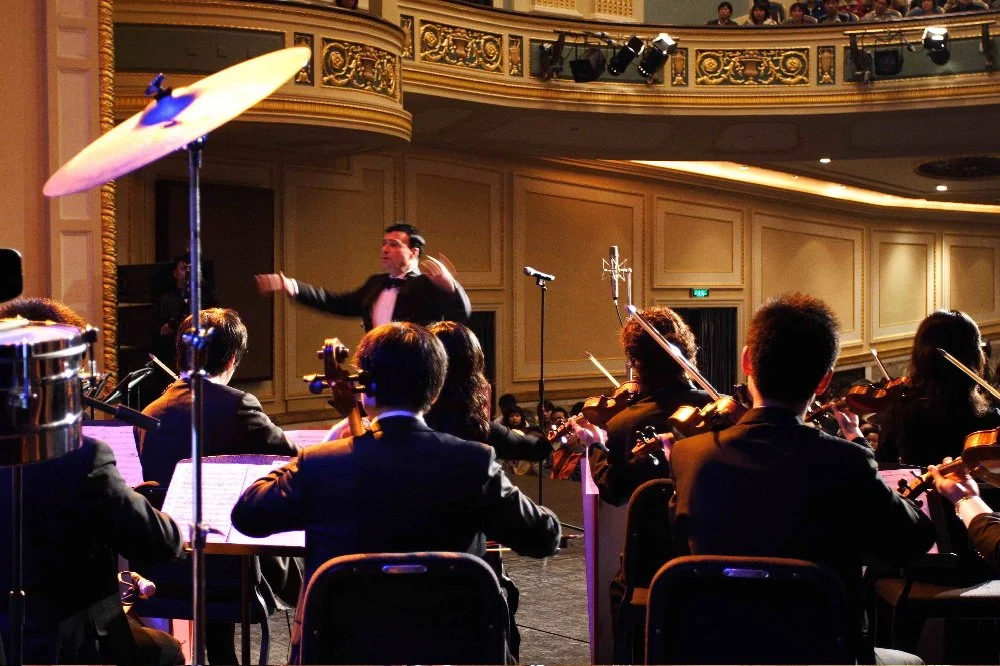 Orchestra performing on stage with conductor leading, in an elegant concert hall with decorative gold accents and balcony seating