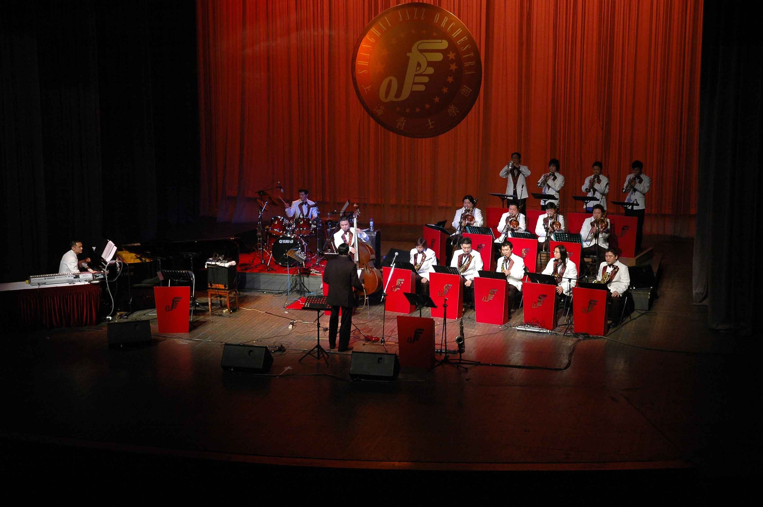 An orchestra performing on stage with a conductor in front. Musicians are playing various instruments including saxophones, drums, and keyboard, dressed in white suits. A large red curtain with a circular logo is in the background.