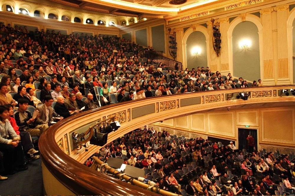 Crowded theater with audience seated in a grand hall, some read programs, ornate decorations and balconies.