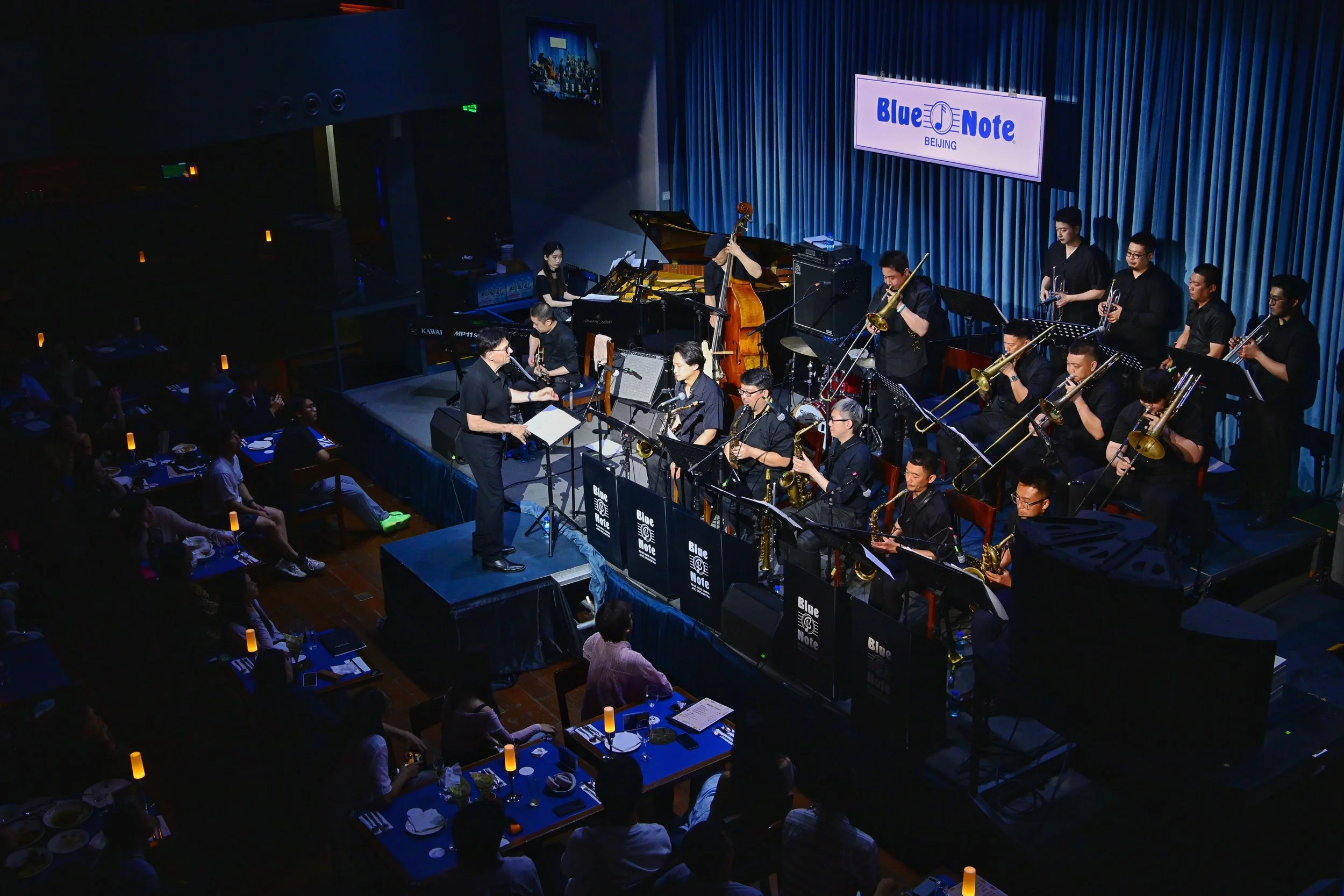 An indoor concert stage with a jazz band performing, featuring musicians playing various instruments such as saxophones, trombones, piano, double bass, and drums. The backdrop has a sign that reads 'Blue Note Beijing,' and there are audience members seated at tables watching the performance.