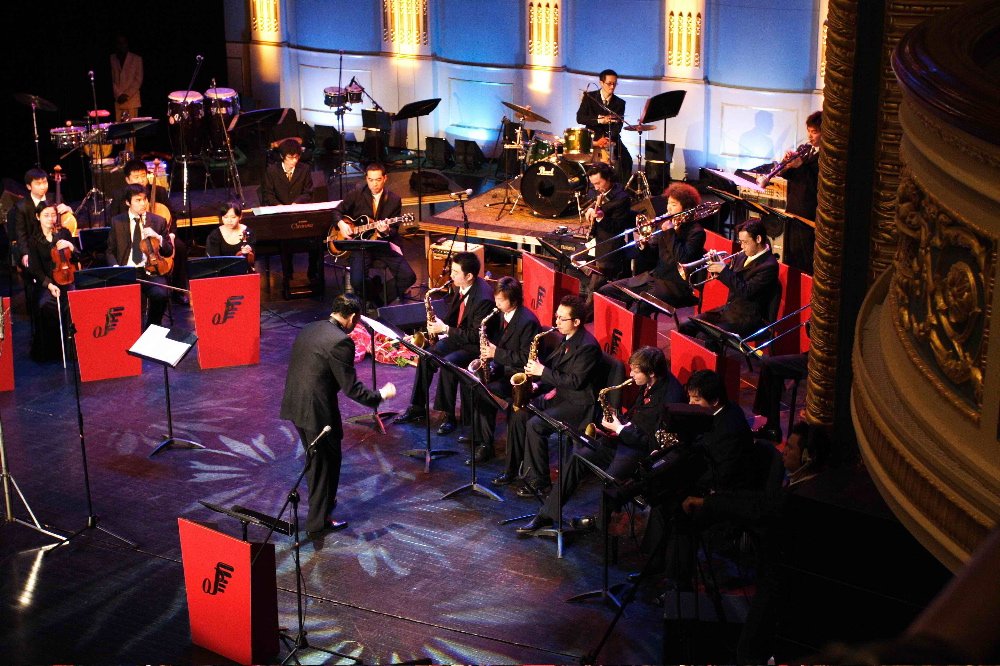 Jazz orchestra on stage with musicians playing violins, guitars, saxophones, trombones, drums, keyboard, and conductor leading the ensemble in a concert hall.