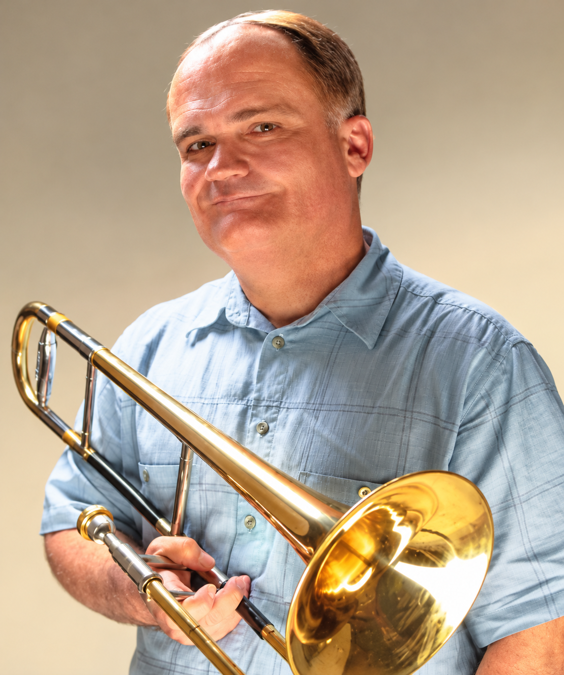 Man smiling while holding a golden trombone, wearing a light blue button-up shirt, against a plain beige background.